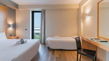 A hotel room with a large bed, a smaller bed, a desk, and a chair. There is a window with curtains, a wall-mounted light, and a view of buildings outside.