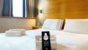 A hotel room with a bed, two pillows, a sign indicating the room has been sanitized, a nightstand with water bottles, and a window with curtains. - B&B HOTEL Pisa Airport (Pisa)