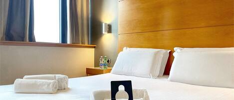 Una habitación de hotel con una cama, dos almohadas, un cartel que indica que la habitación ha sido desinfectada, una mesita de noche con botellas de agua y una ventana con cortinas.