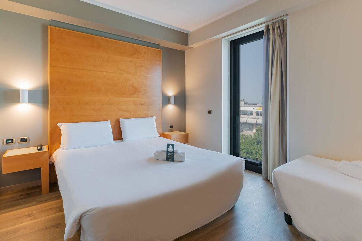 A hotel room with a large bed, a smaller bed, wooden headboard, bedside lamps, a nightstand, and a view of the city through the window.