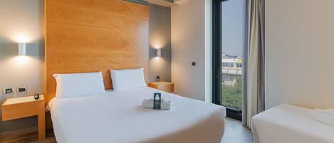 Een hotelkamer met een groot bed, een kleiner bed, een houten hoofdbord, nachtlampjes, een nachtkastje en uitzicht op de stad door het raam.