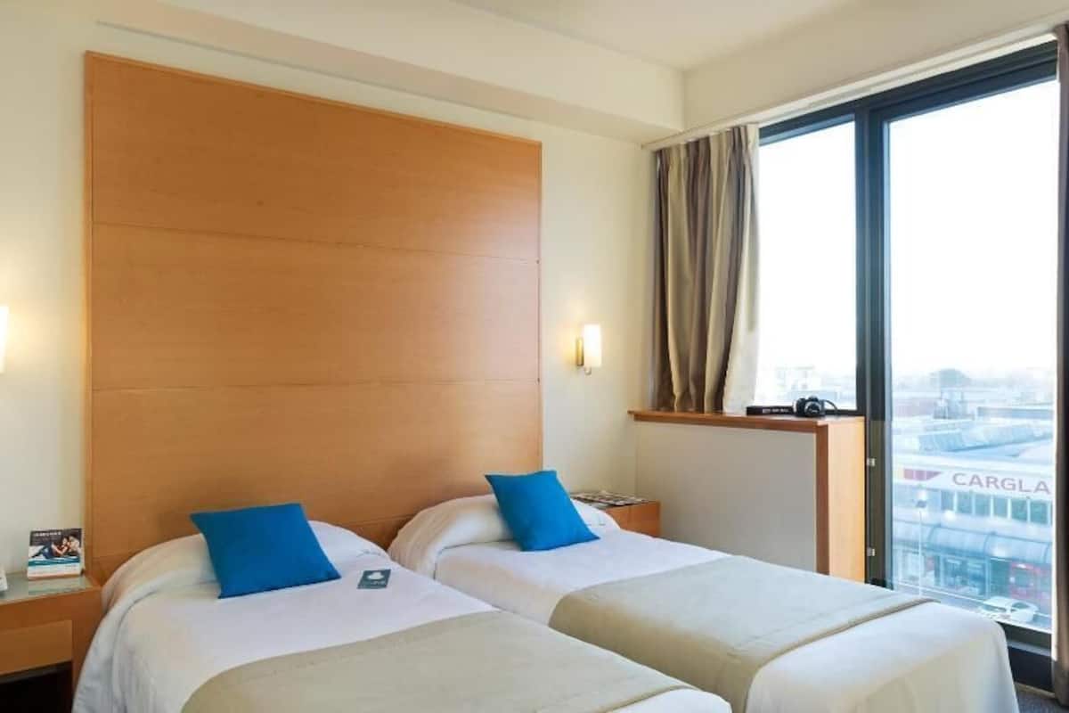 A hotel room with two beds, a wooden headboard, a nightstand with a phone, and a window with a view of a bridge.
