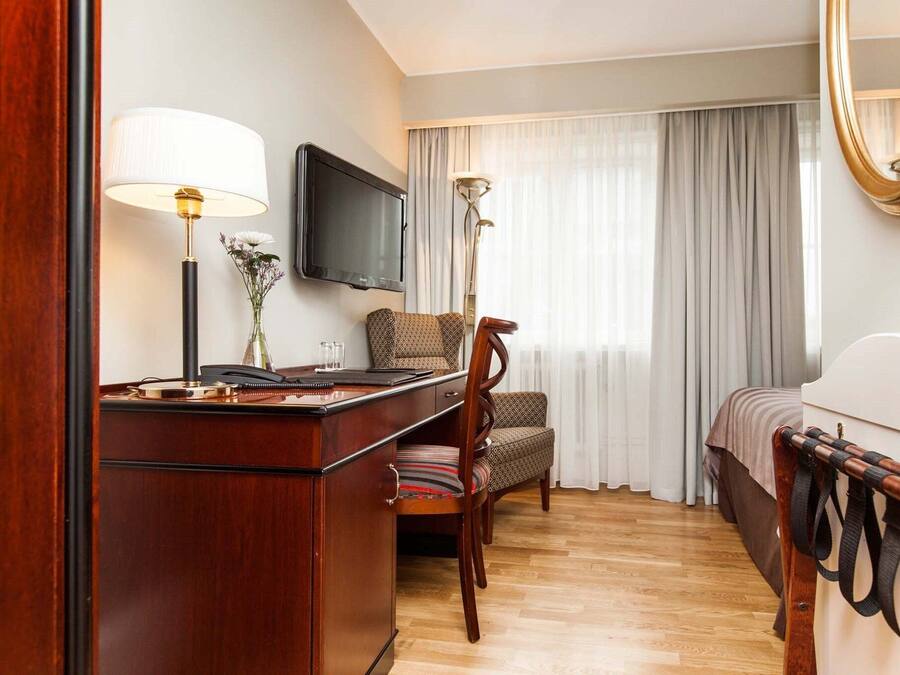 Een hotelkamer met een houten bureau, een stoel, een bed, een aan de muur bevestigde televisie en een groot raam met gordijnen.