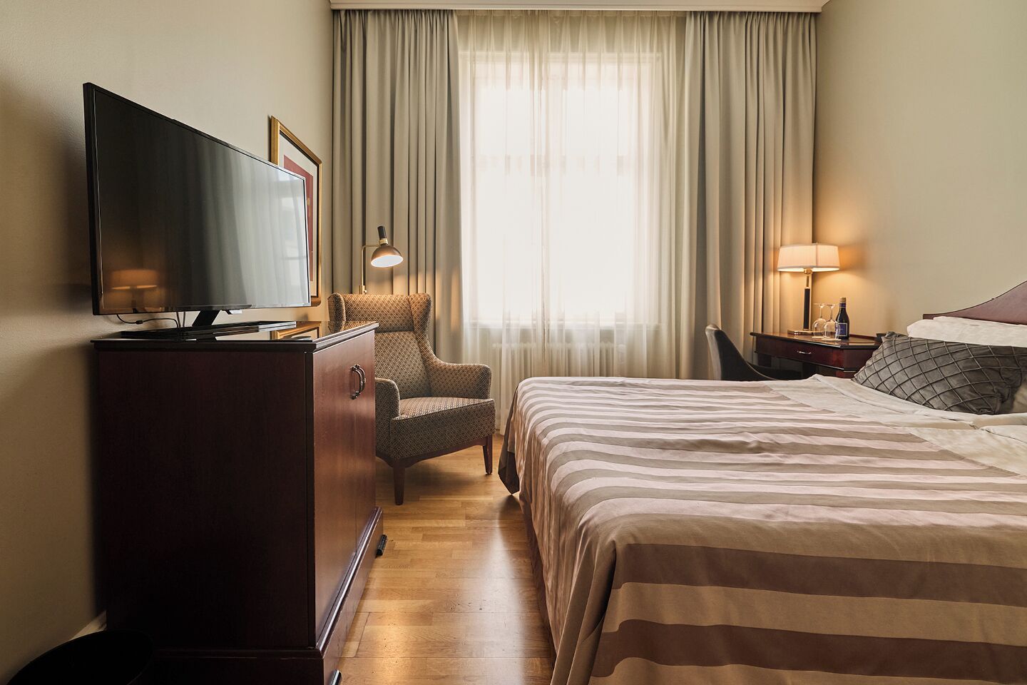 A hotel room with a bed, a TV, a chair, a desk with a lamp, and a window with curtains.