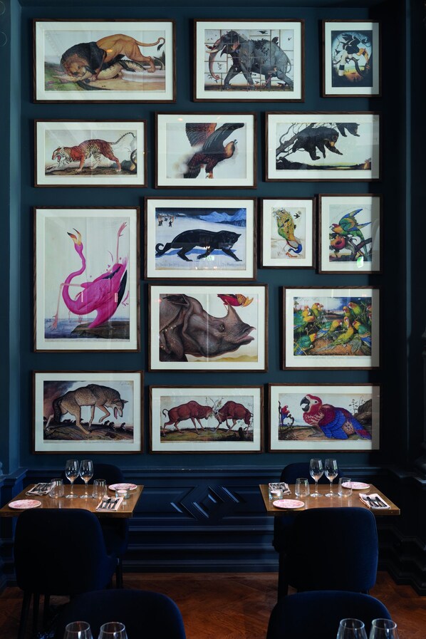 Un comedor con una pared de cuadros de animales, mesas dispuestas con copas de vino y sillas oscuras azul.