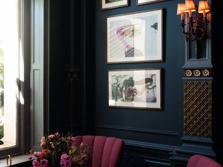 A room with a dark wall, a window, a round table with a vase of flowers, and a red upholstered chair.
