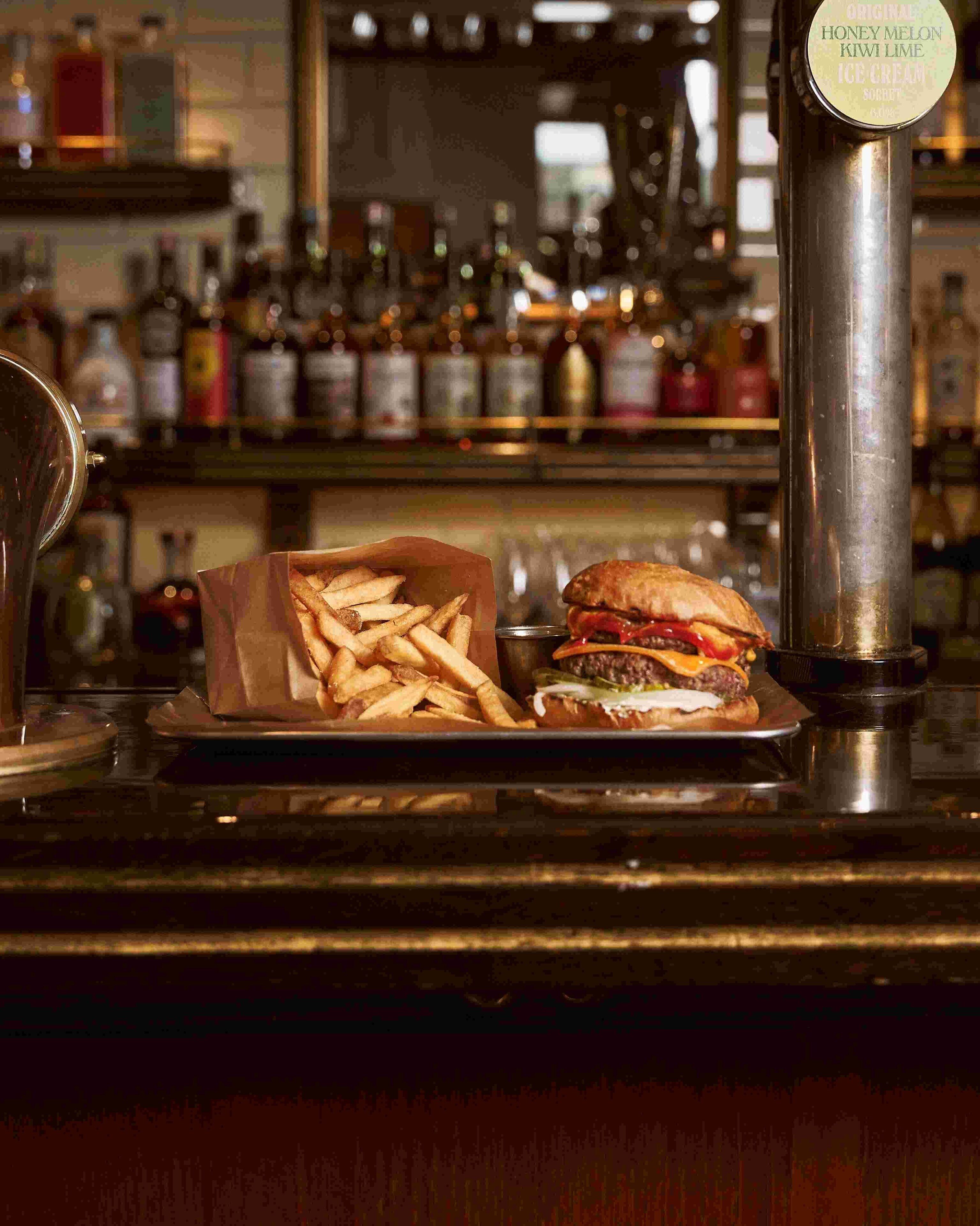 Una barra con una hamburguesa y papas fritas, un grifo de cerveza y estantes con botellas al fondo.