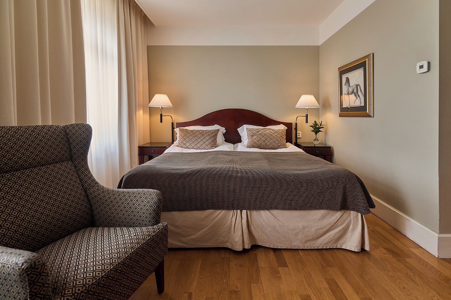 A hotel room with a large bed, two bedside lamps, a nightstand, a chair, and a framed picture on the wall.
