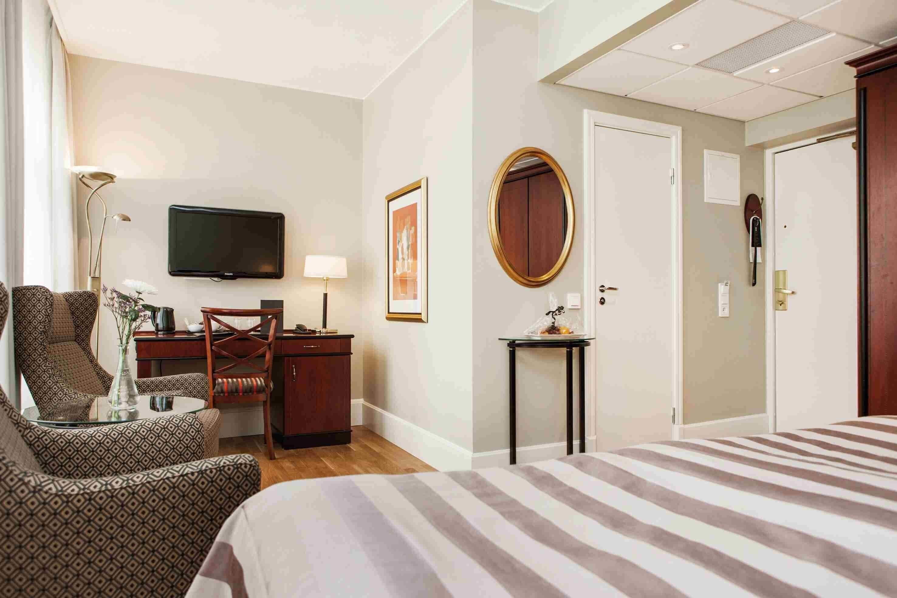 Ein Hotelzimmer mit einem Bett, einem Schreibtisch mit Stuhl, einem Fernseher und einem kleinen Tisch mit Lampe.
