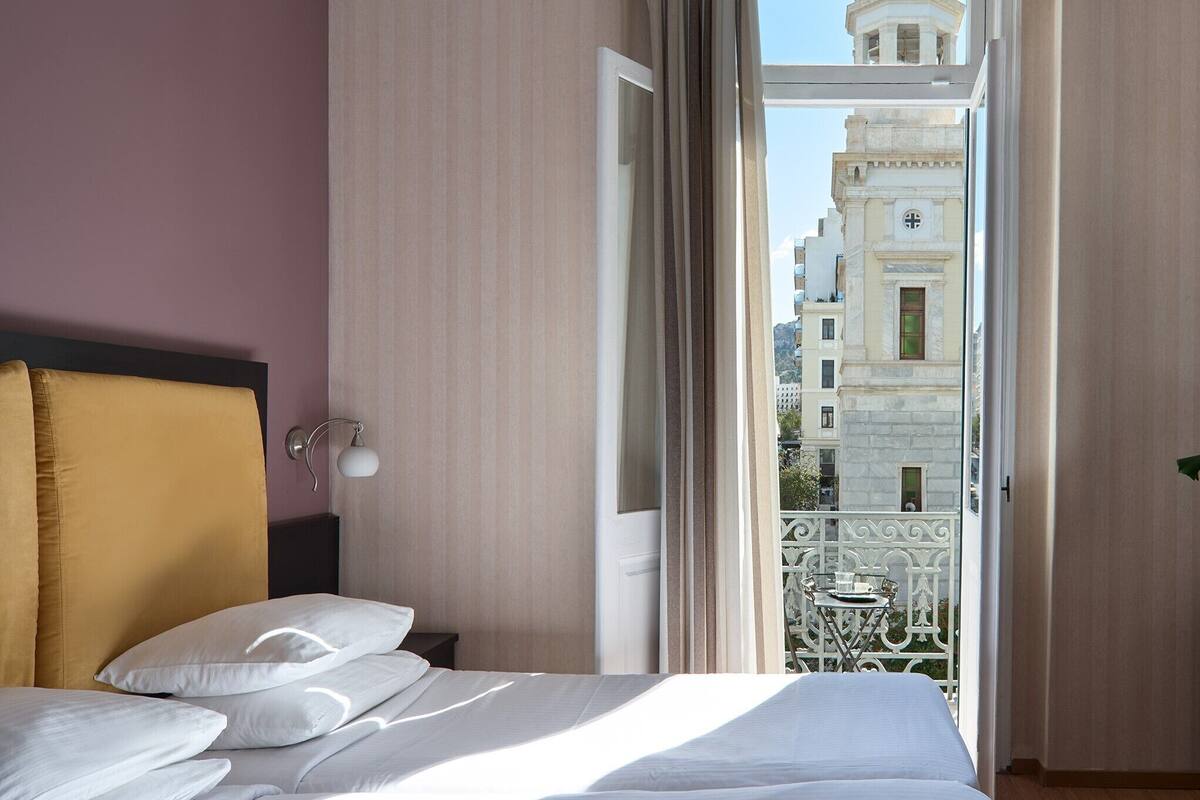 A hotel room with a bed, a nightstand, a lamp, and a view of a building through an open door.