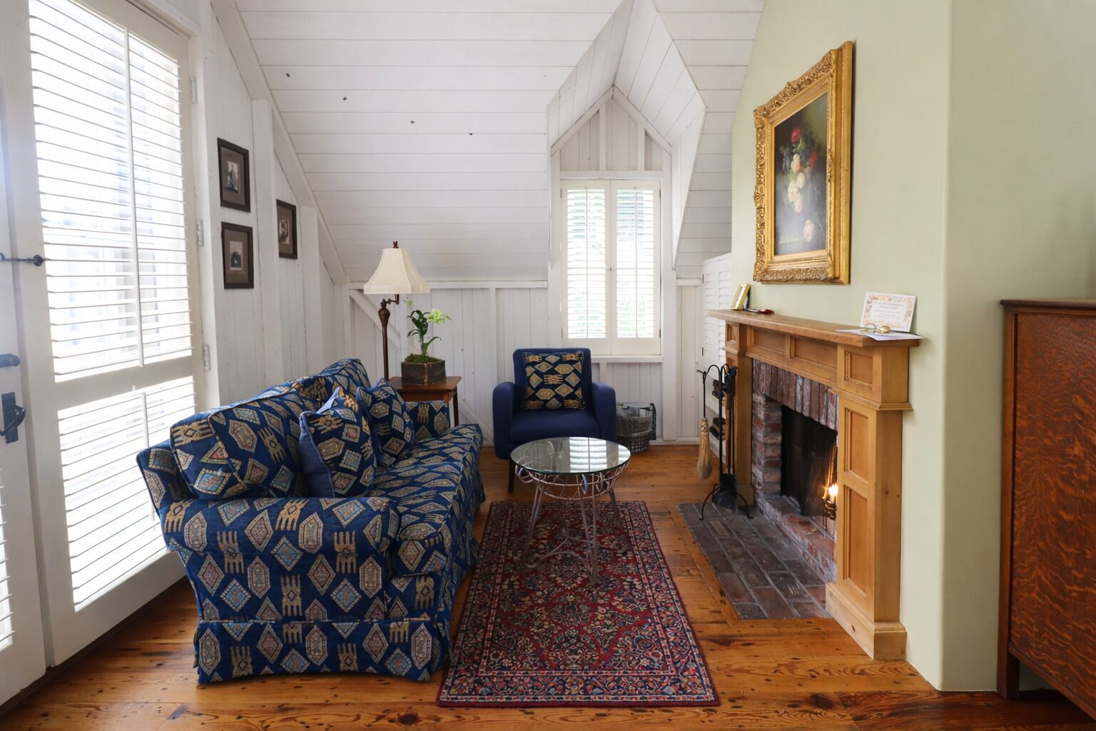 Ein Zimmer mit einem blauen, gemusterten Sofa, einem runden Glastisch, einem roten Teppich mit Muster und einem Kamin. An der Wand hängt ein Gemälde.
