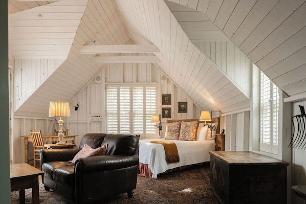 Een kamer met een schuine plafond, een bed met een witte sprei, een leren bank, een houten tafel en een lamp.