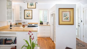 A modern kitchen with white cabinetry, a wooden countertop, and a central island. There are framed botanical prints on the walls, a potted orchid on the island, and a dining area with a red upholstered chair.
