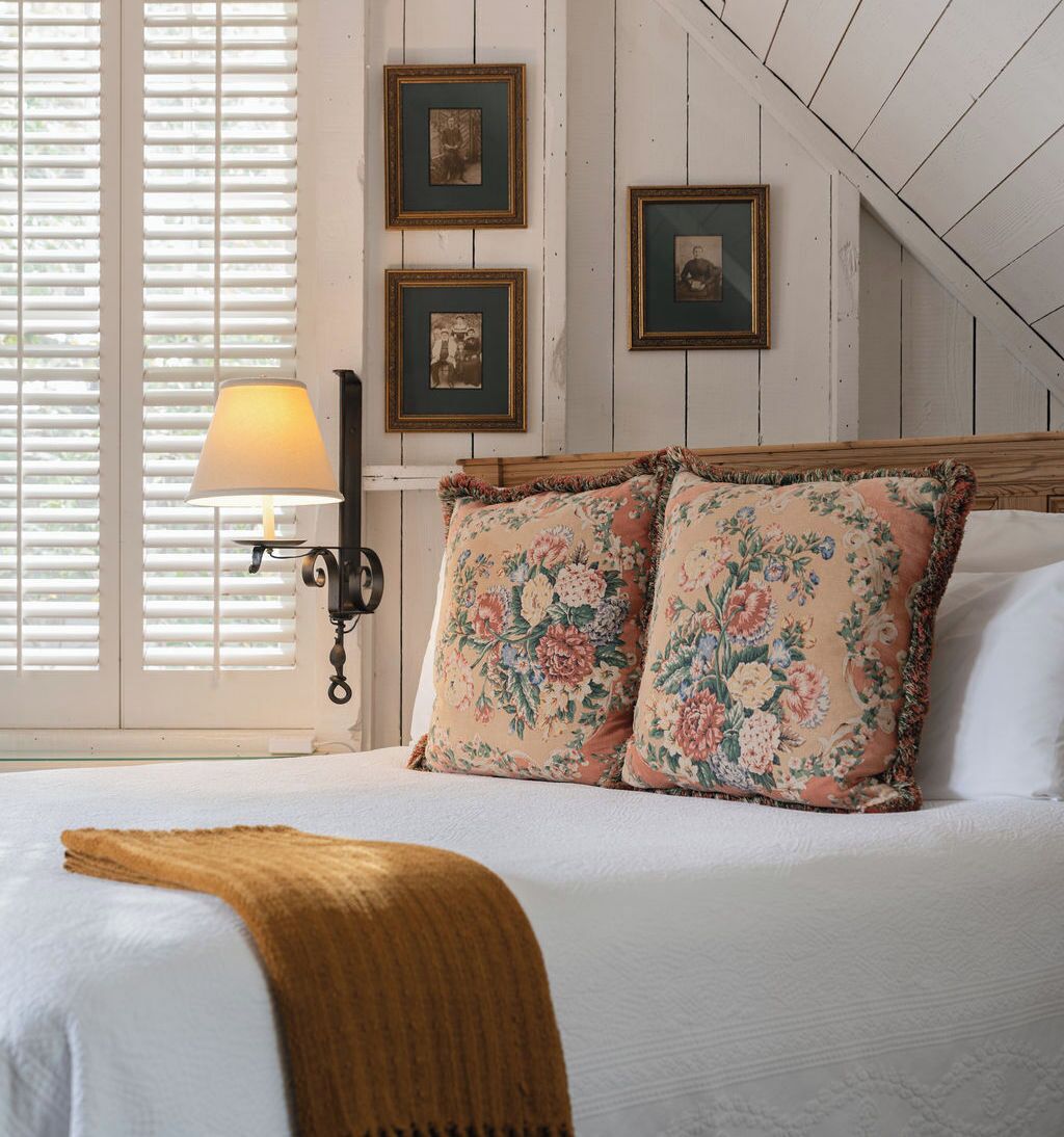 Un dormitorio con cabecera de madera, dos almohadas estampadas, una lámpara de noche y cuadros en la pared.