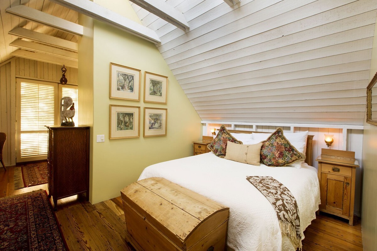 A bedroom with a sloped ceiling, a bed with floral pillows, a wooden chest, a dresser, and framed pictures on the wall.