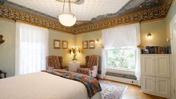 A bedroom with a large bed, two chairs, a small table with a lamp, a window with curtains, and a bookshelf.