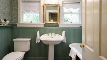 A bathroom with a white toilet, a pedestal sink, and a bathtub. There are two windows with decorative shutters, and a mirror above the sink.