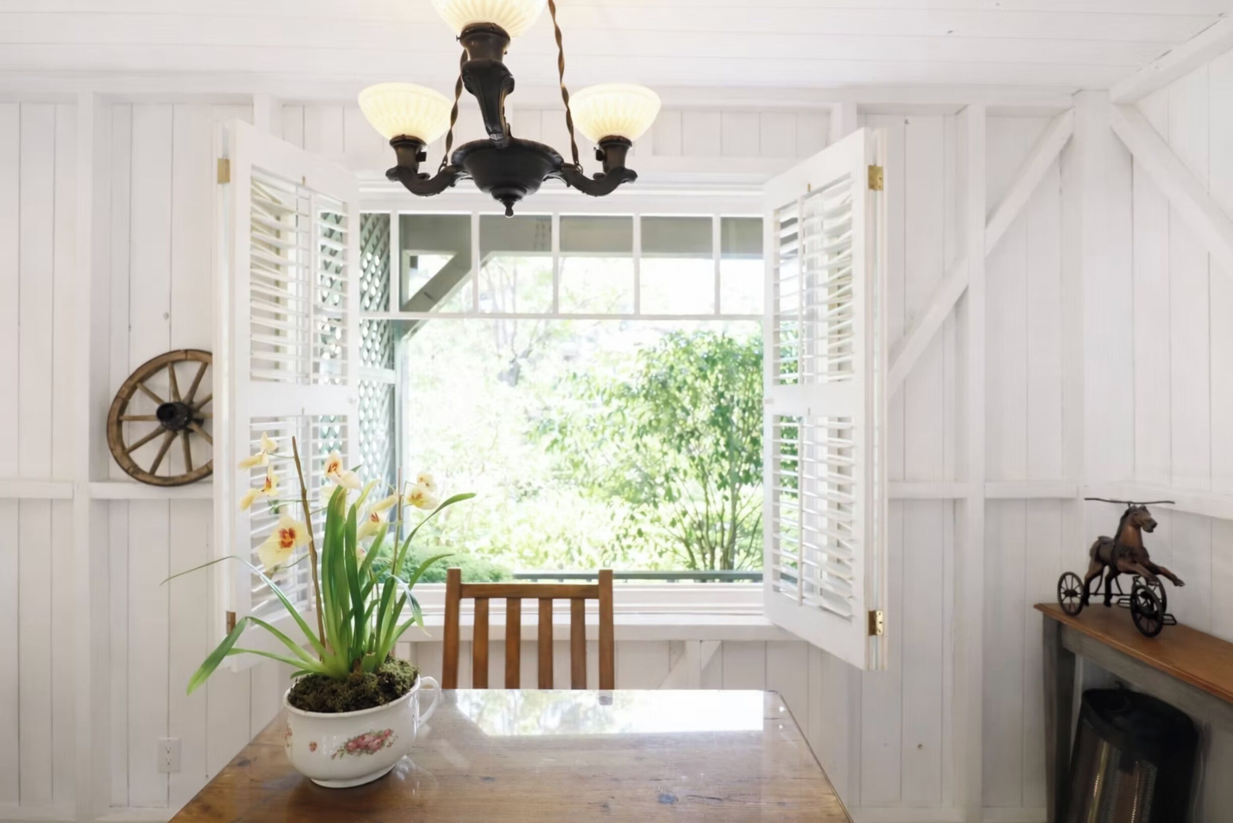 A dining area with a table, chairs, a chandelier, a window with shutters, and a decorative wheel.
