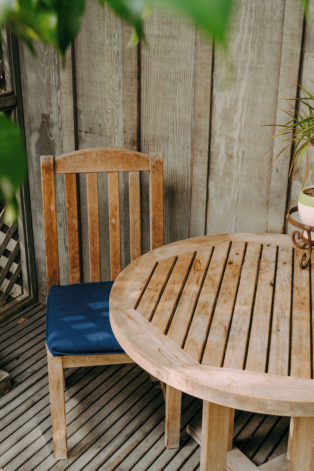 A wooden outdoor seating area with a round table and a single wooden chair with a blue cushion.