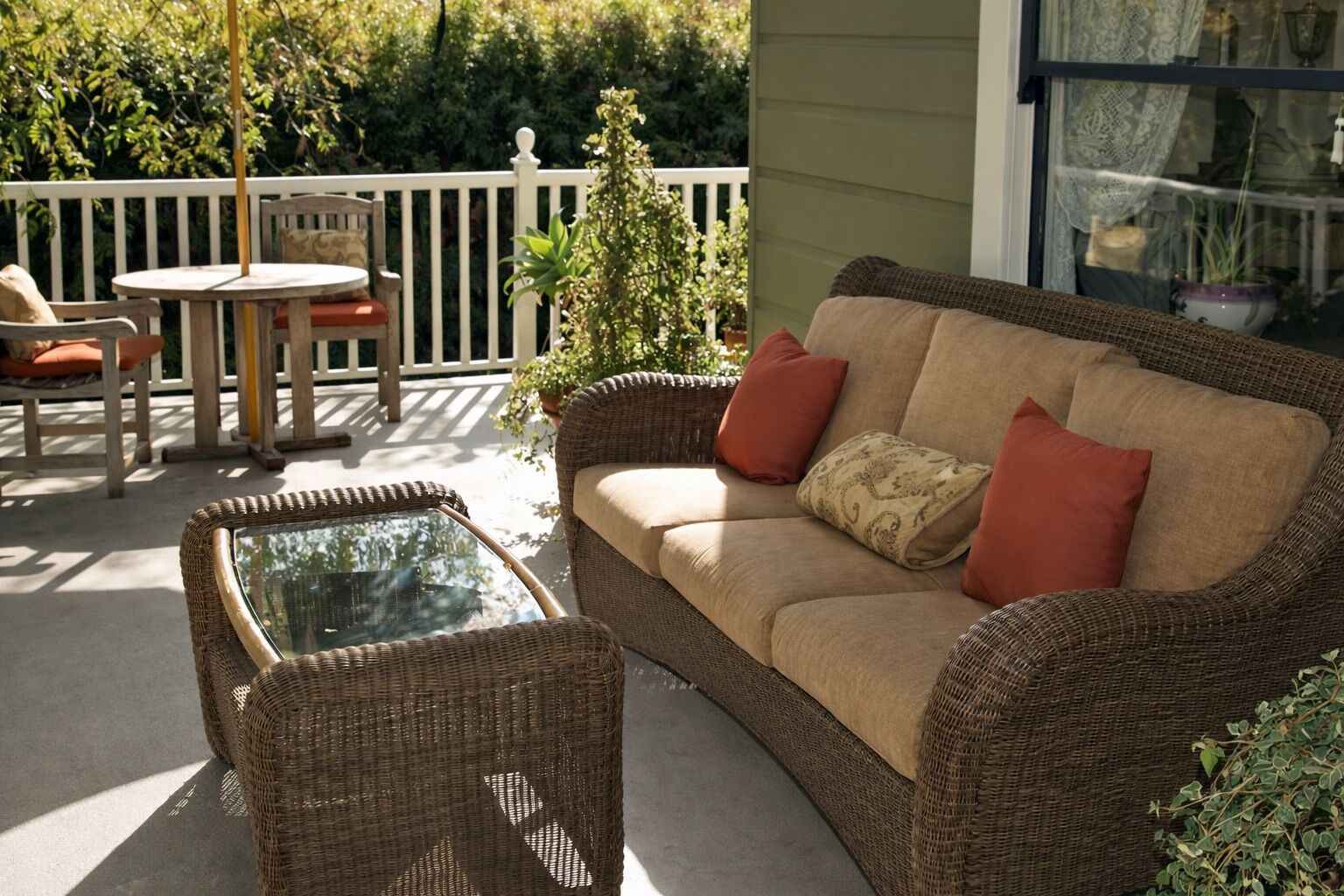 Un sofá de mimbre con cojines, una mesa con cubierta de vidrio y una silla en un patio exterior con baranda blanca.