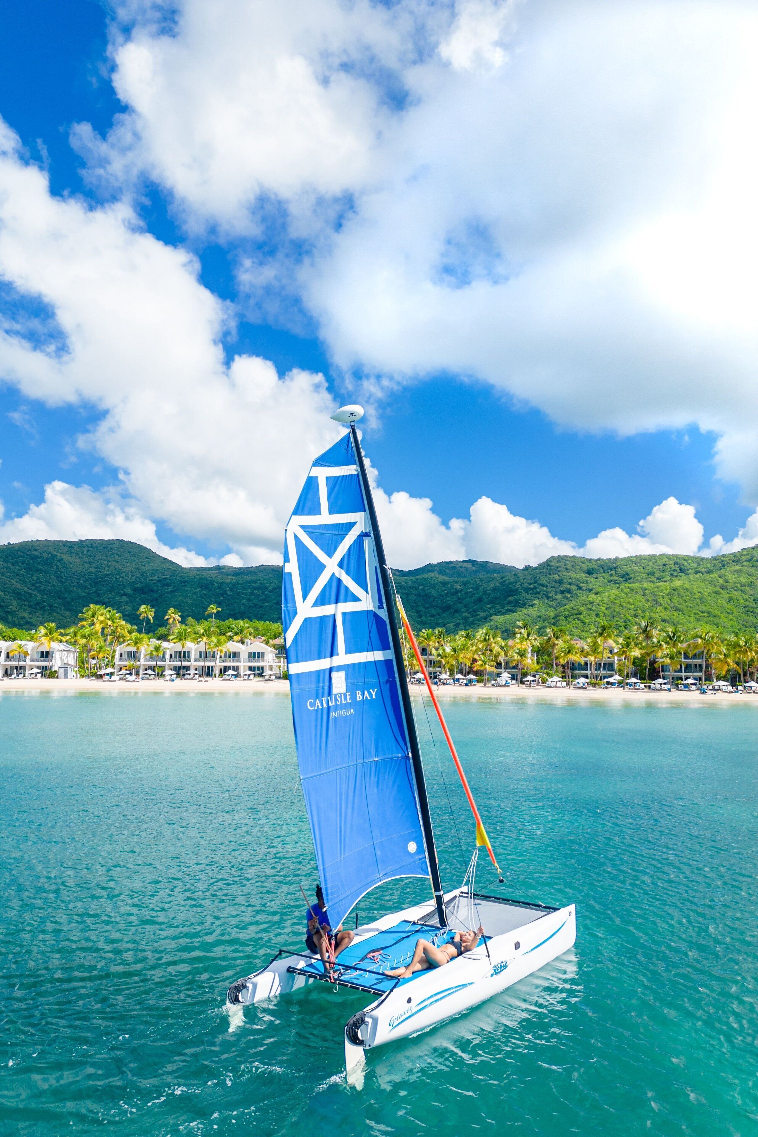 A sailboat with a blue sail on turquoise water, surrounded by a tropical beach and lush greenery.