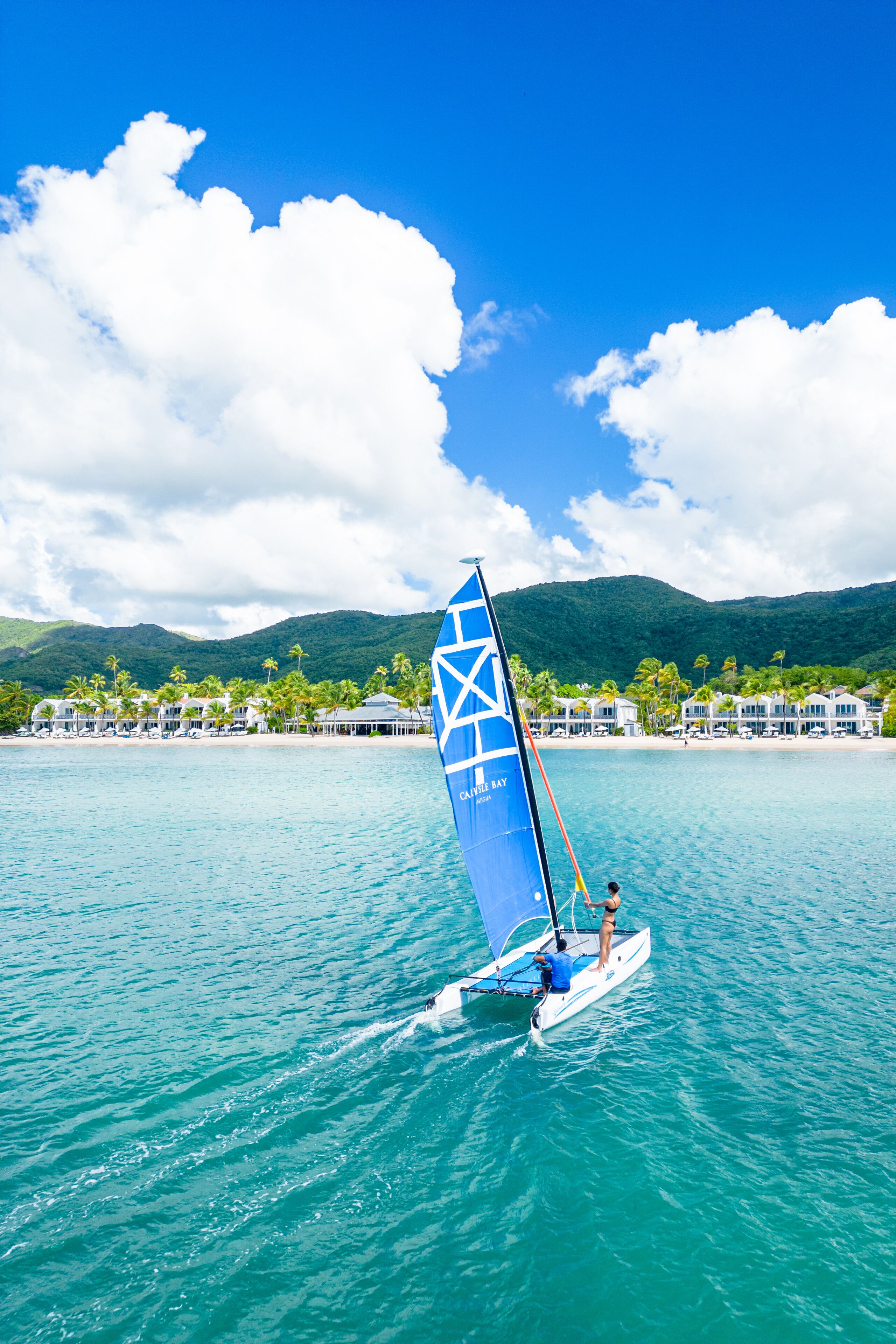 A sailboat with a blue sail on turquoise water, a beach with buildings and palm trees in the background, and mountains in the distance.