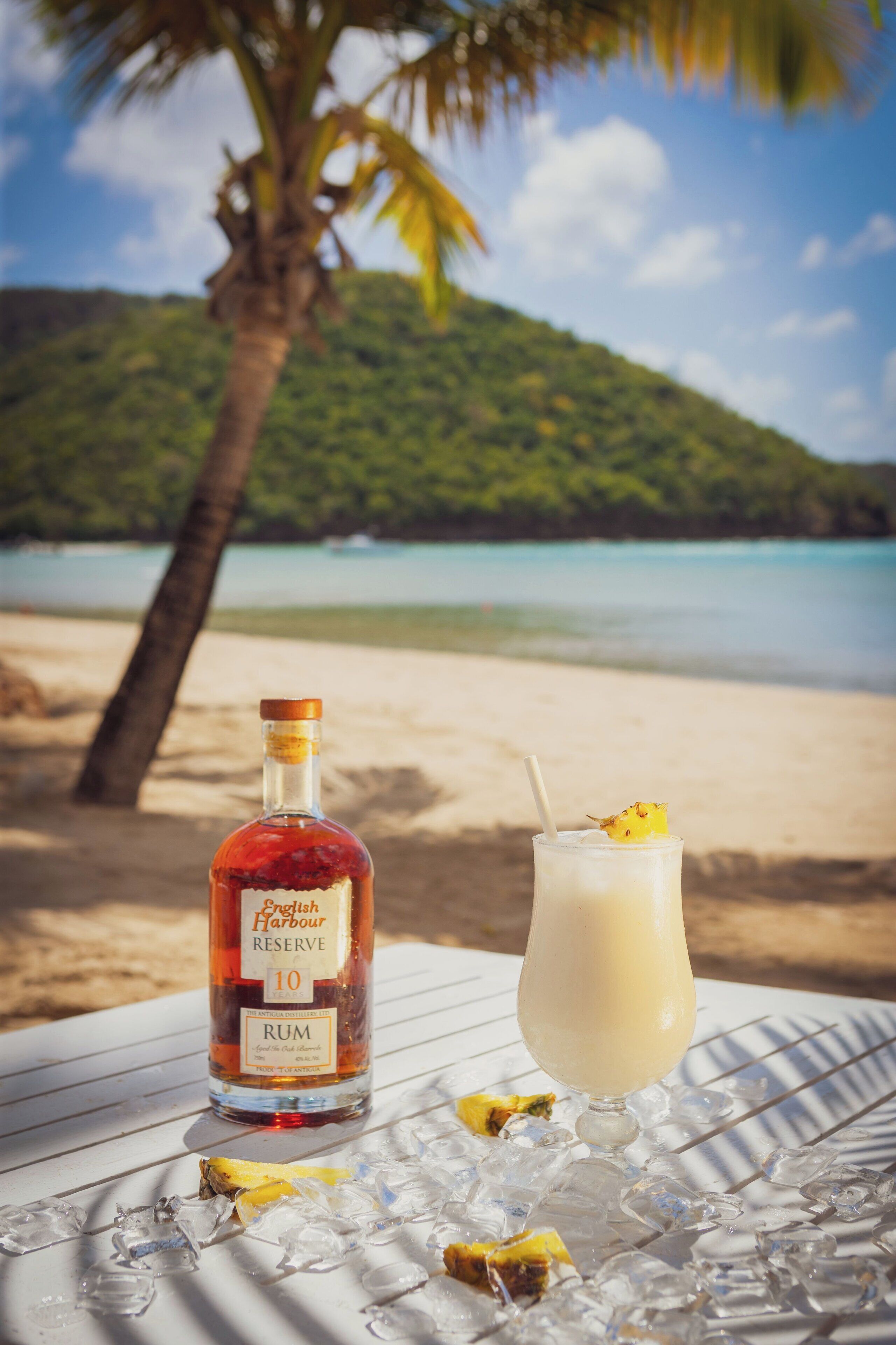 A bottle of English Harbour Reserve Rum and a glass of a light-colored beverage with a straw, ice, and a pineapple slice on a table with a beach and palm tree in the background.
