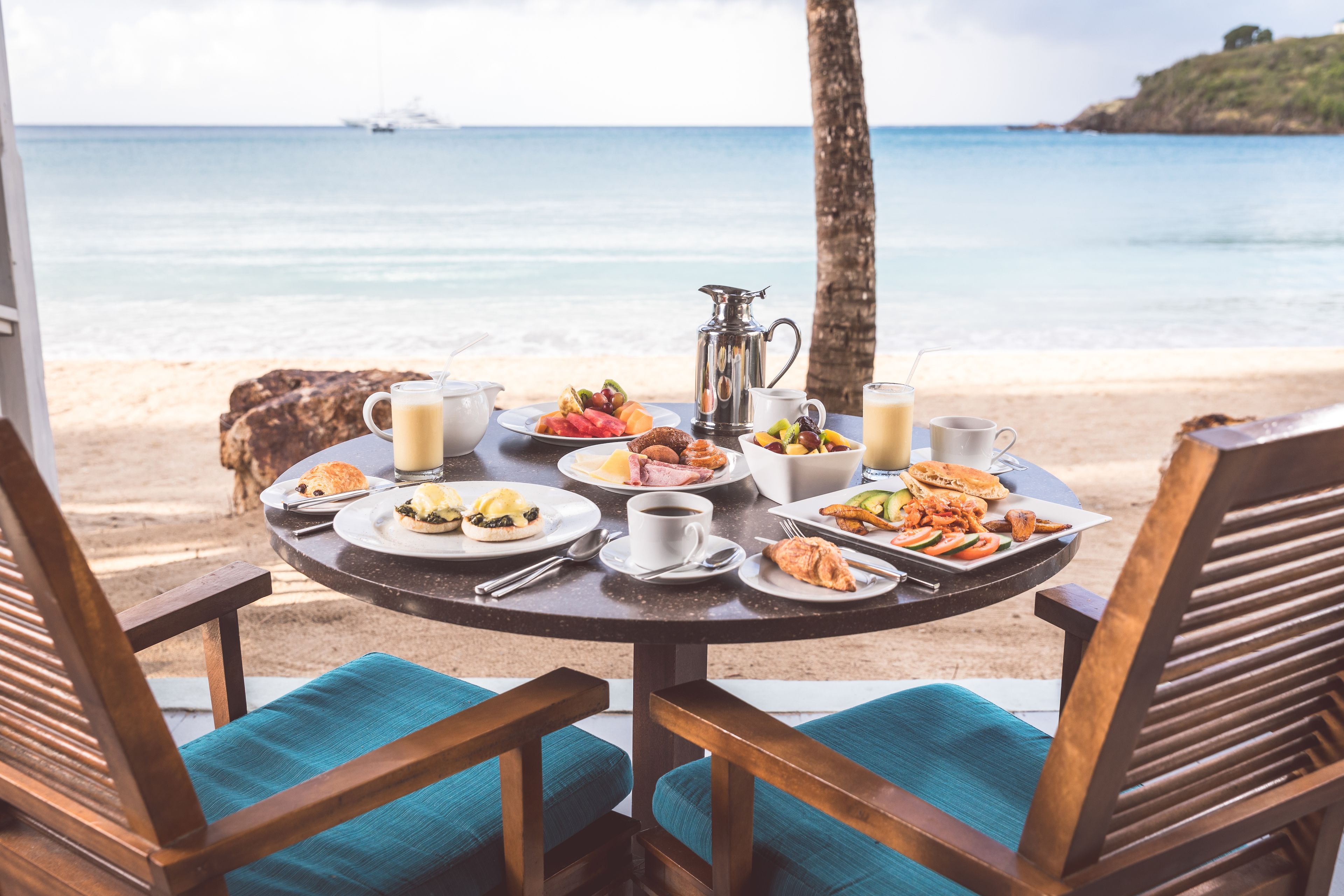 Et rundt bord med morgenmadsretter og en kaffekop, med udsigt over en strand med et skib i det fjerne.