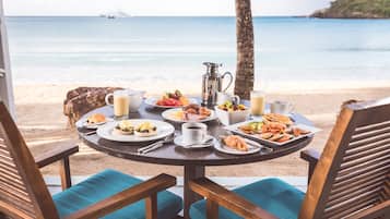 Et rundt bord med morgenmadsretter og en kaffekop, med udsigt over en strand med et skib i det fjerne.