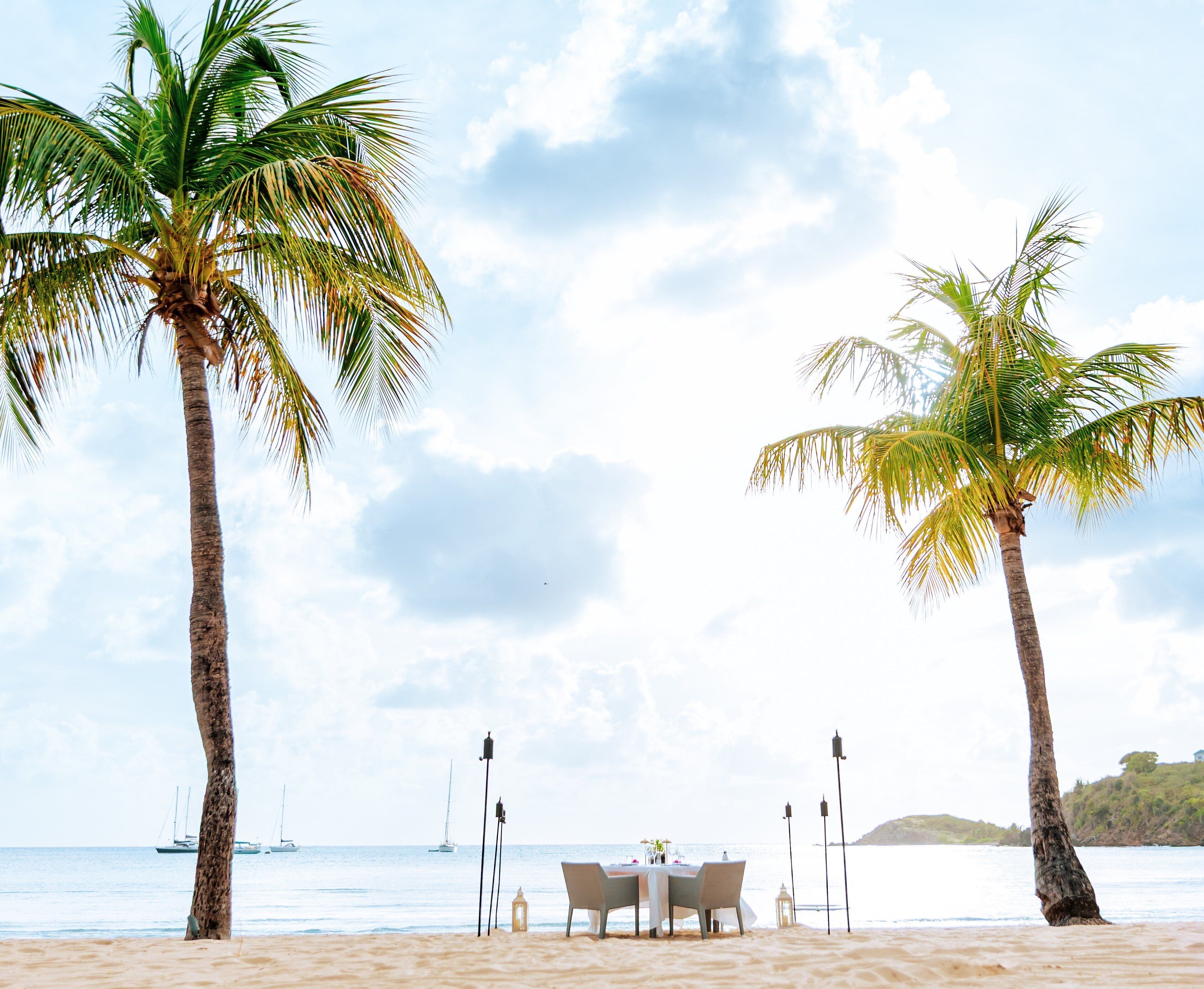 A beachside dining setup with a table and chairs, palm trees, and a view of the sea and boats.
