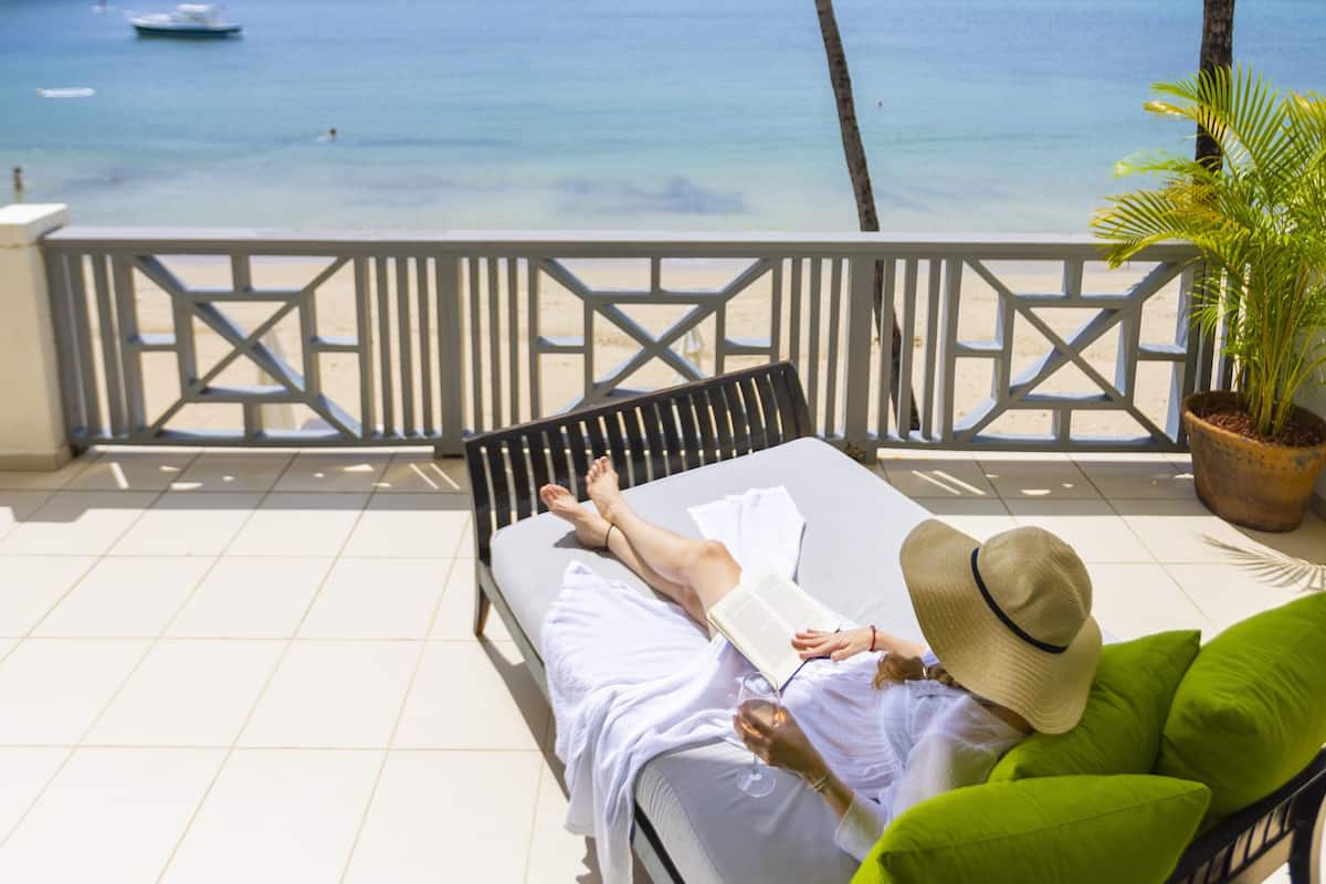 A balcony with a view of the ocean, a palm tree, and a person relaxing on a bed with a book and glass.