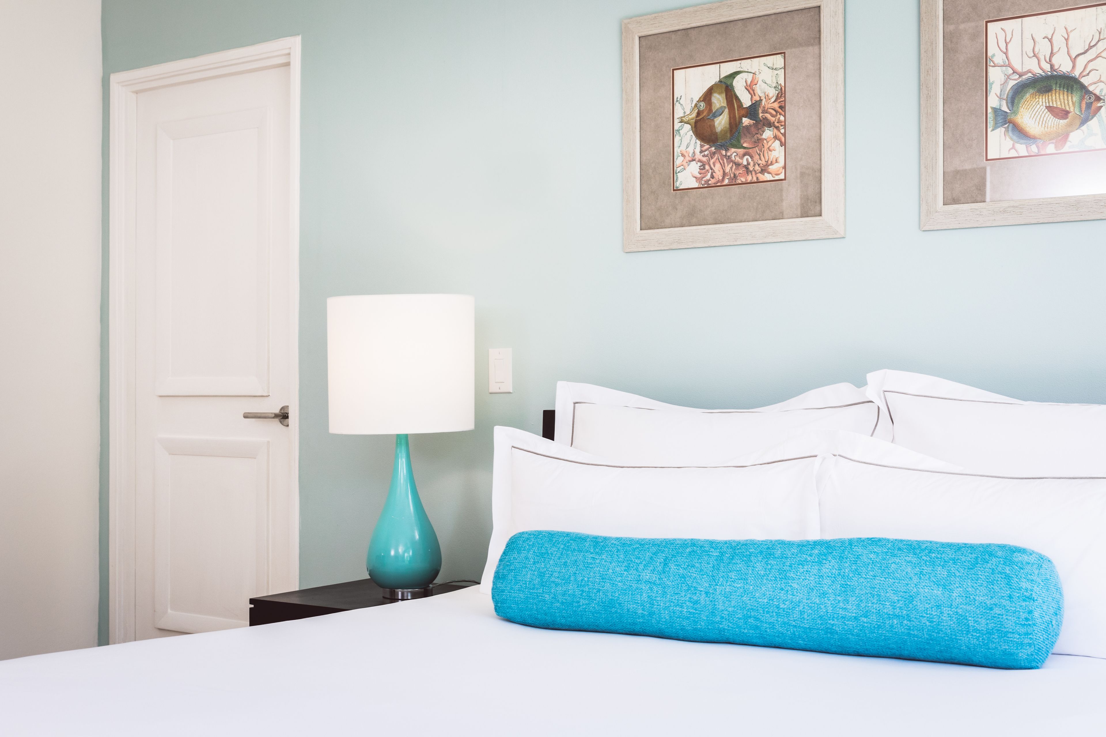 Een opgemaakt bed met witte lakens en een blauw accentkussen, een blauwgroene lamp op een nachtkastje, en twee ingelijste kunstwerken aan de muur.
