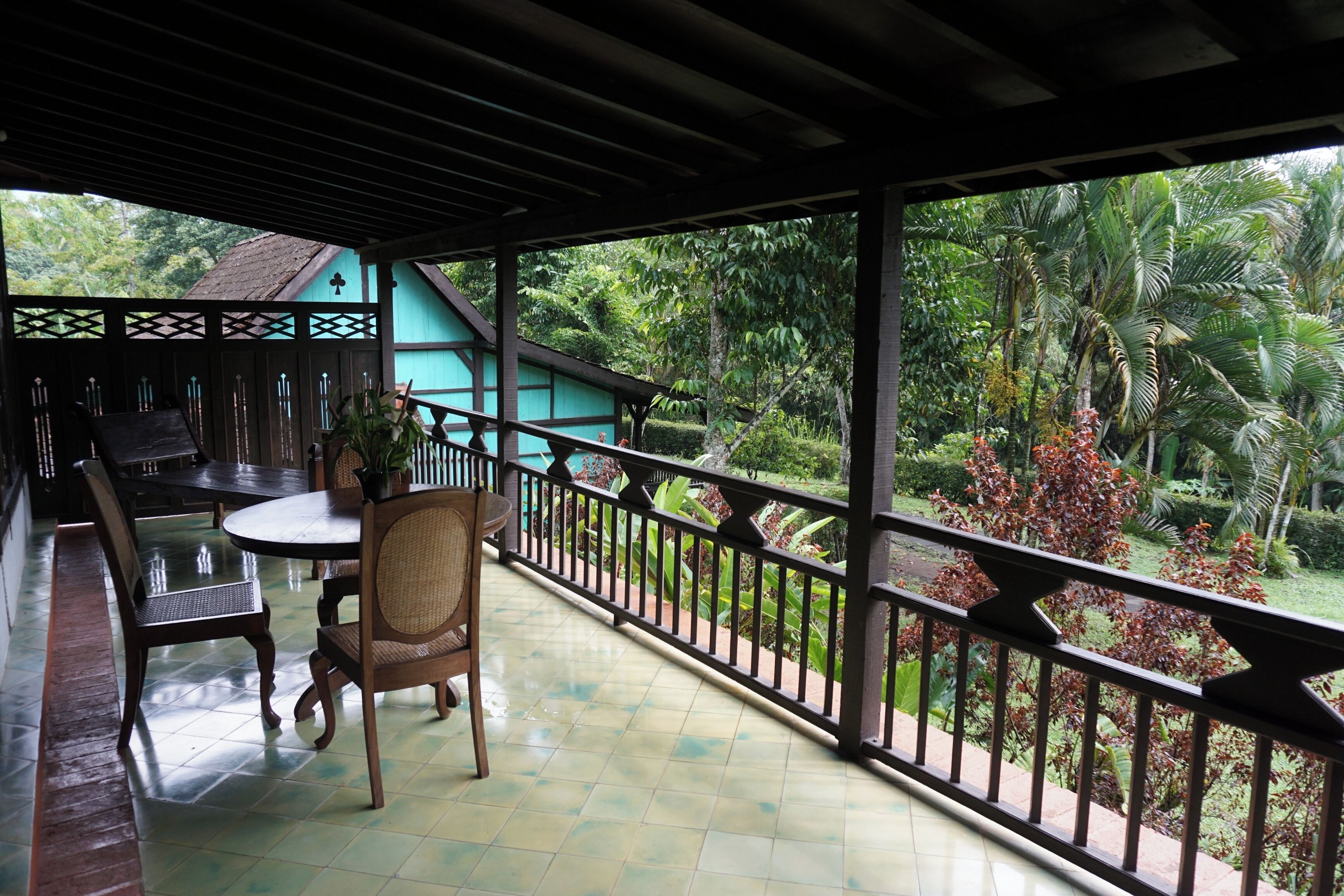 Un balcon abrité, doté d’une table et de chaises, offrant une vue sur une végétation luxuriante, et un bâtiment se distinguant par son toit bleu.