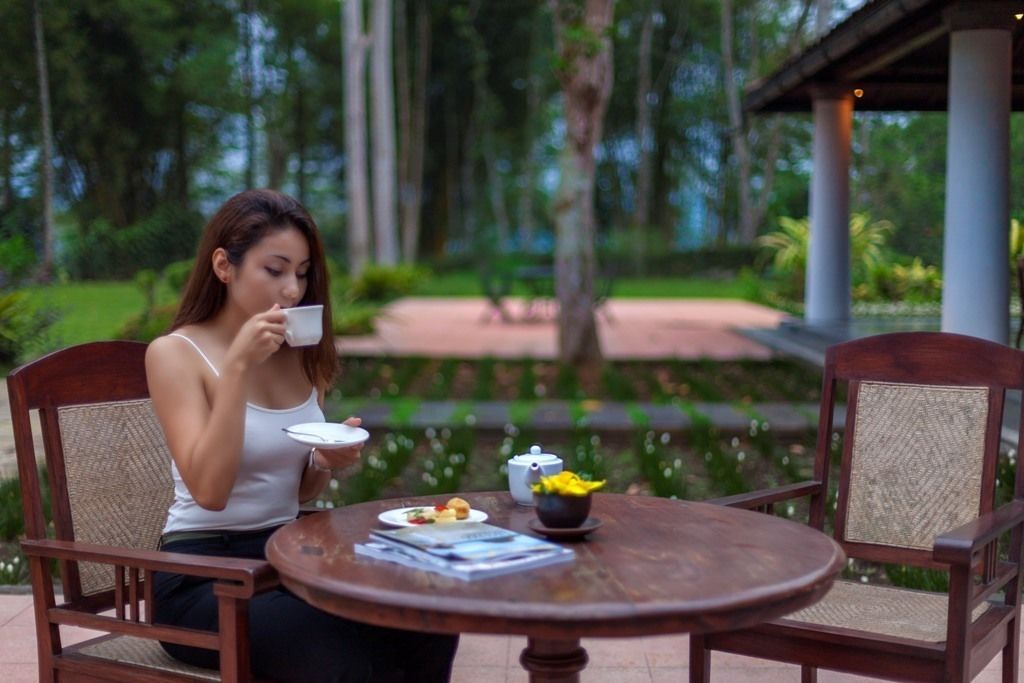 A woman sitting at a round wooden table outdoors, with a cup and a bowl of food on the table.