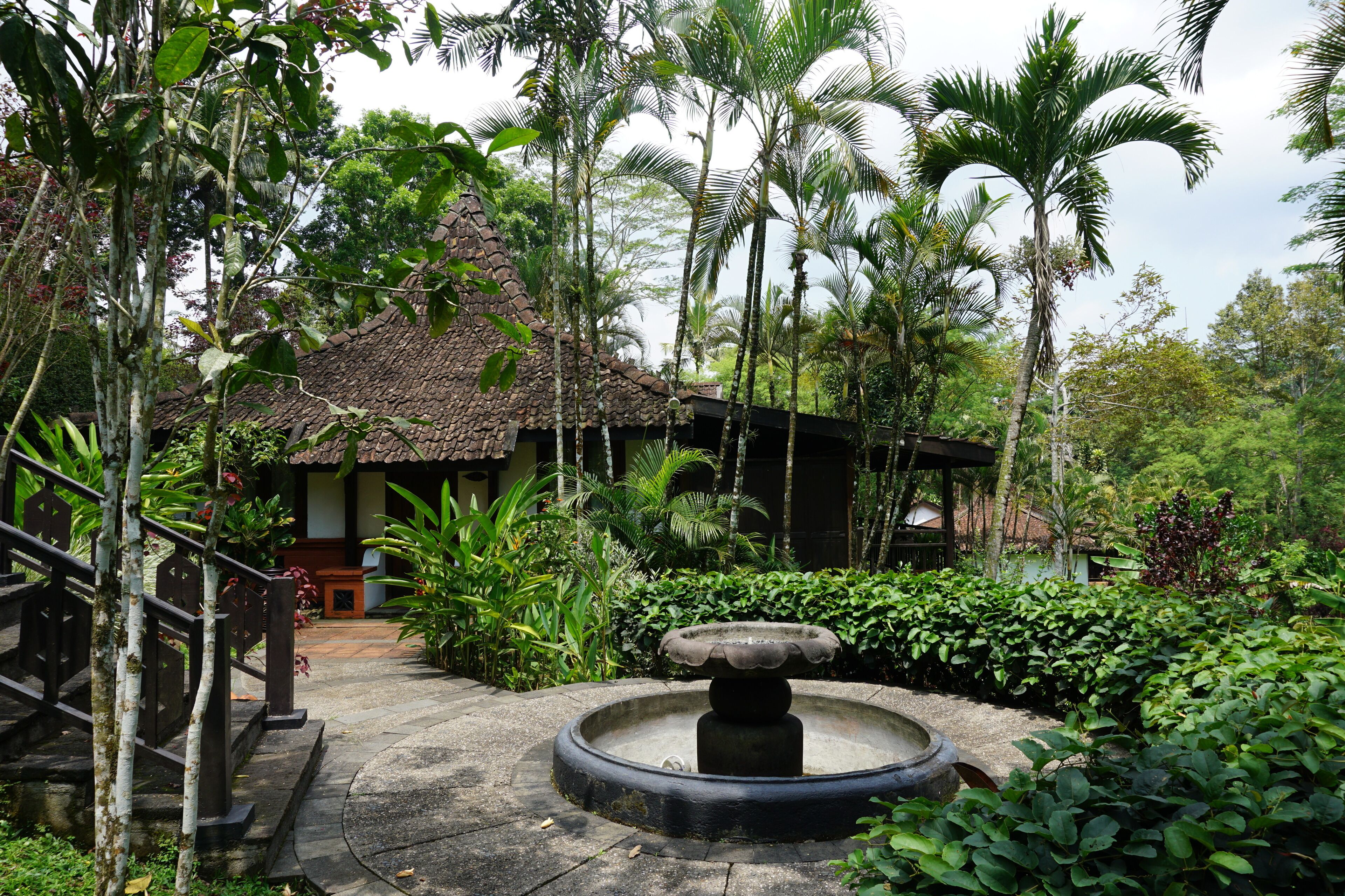 A traditional house with a thatched roof, surrounded by lush greenery and a stone pathway leading to a central fountain.