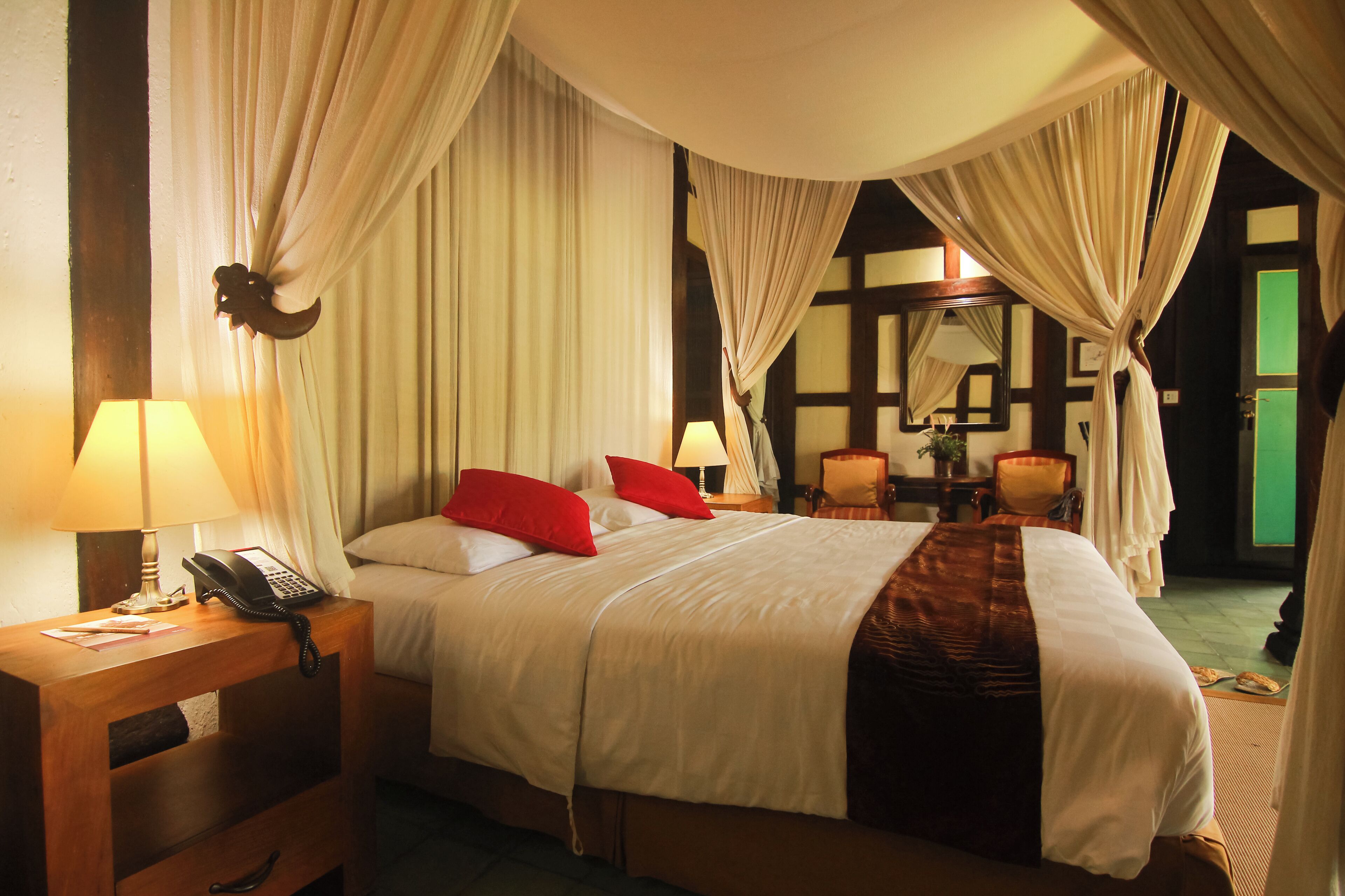 A four-poster bed with white linens and red pillows, a bedside telephone, and a lamp on a wooden nightstand.