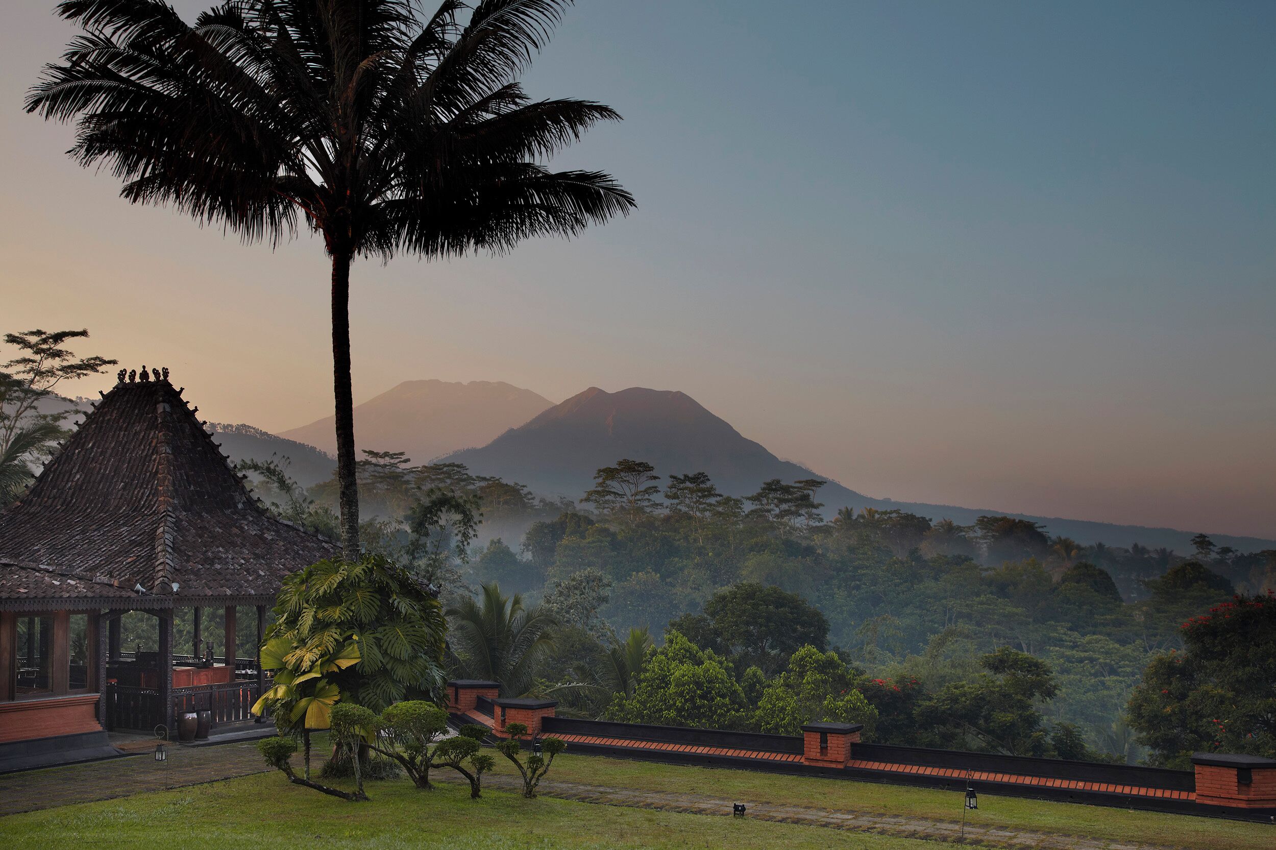 A traditional wooden structure with a thatched roof, surrounded by lush greenery and a view of distant mountains.