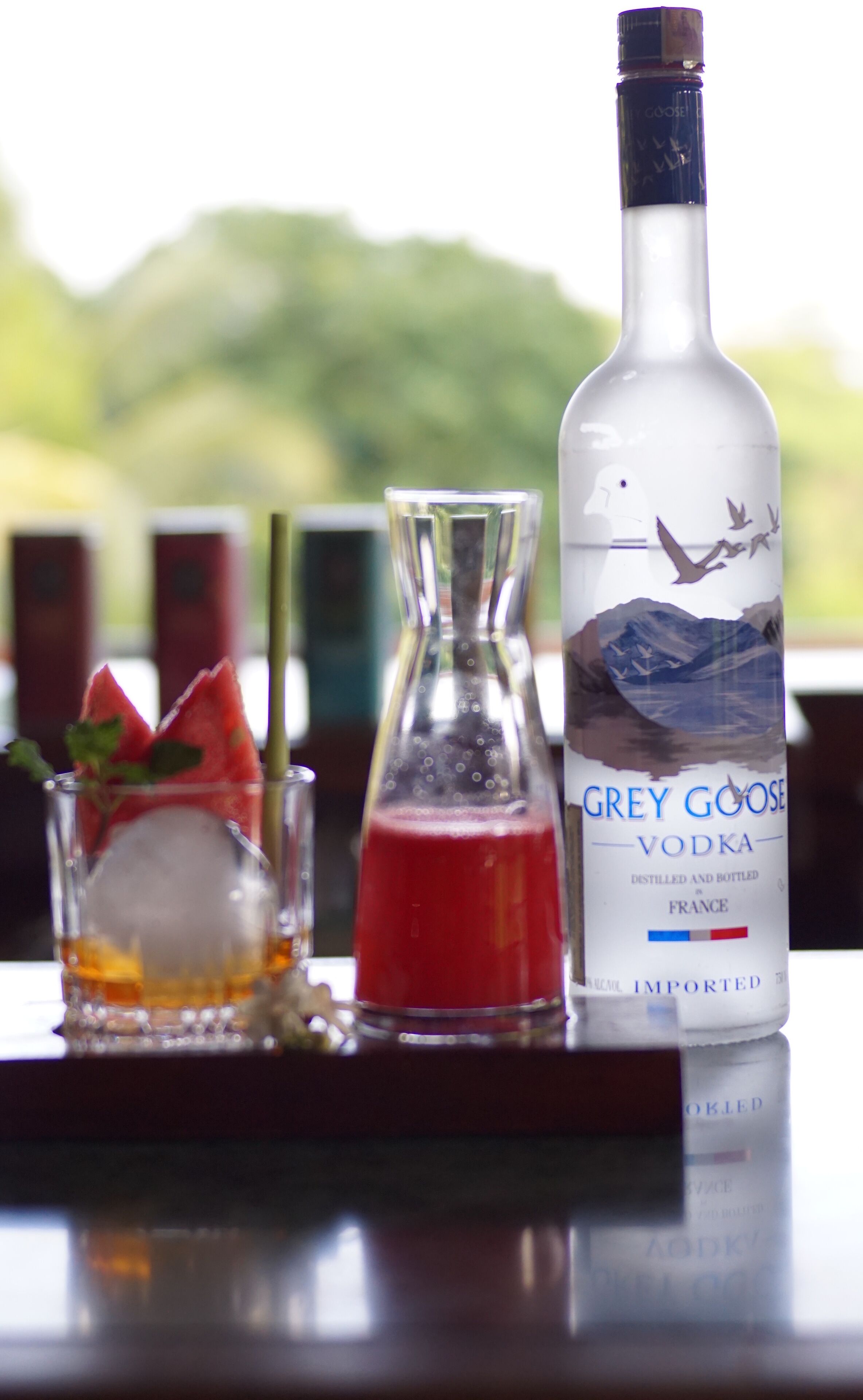 Una botella de vodka Grey Goose, un vaso con una bebida roja, y un vaso con hielo y una tajada de sandía sobre una barra.