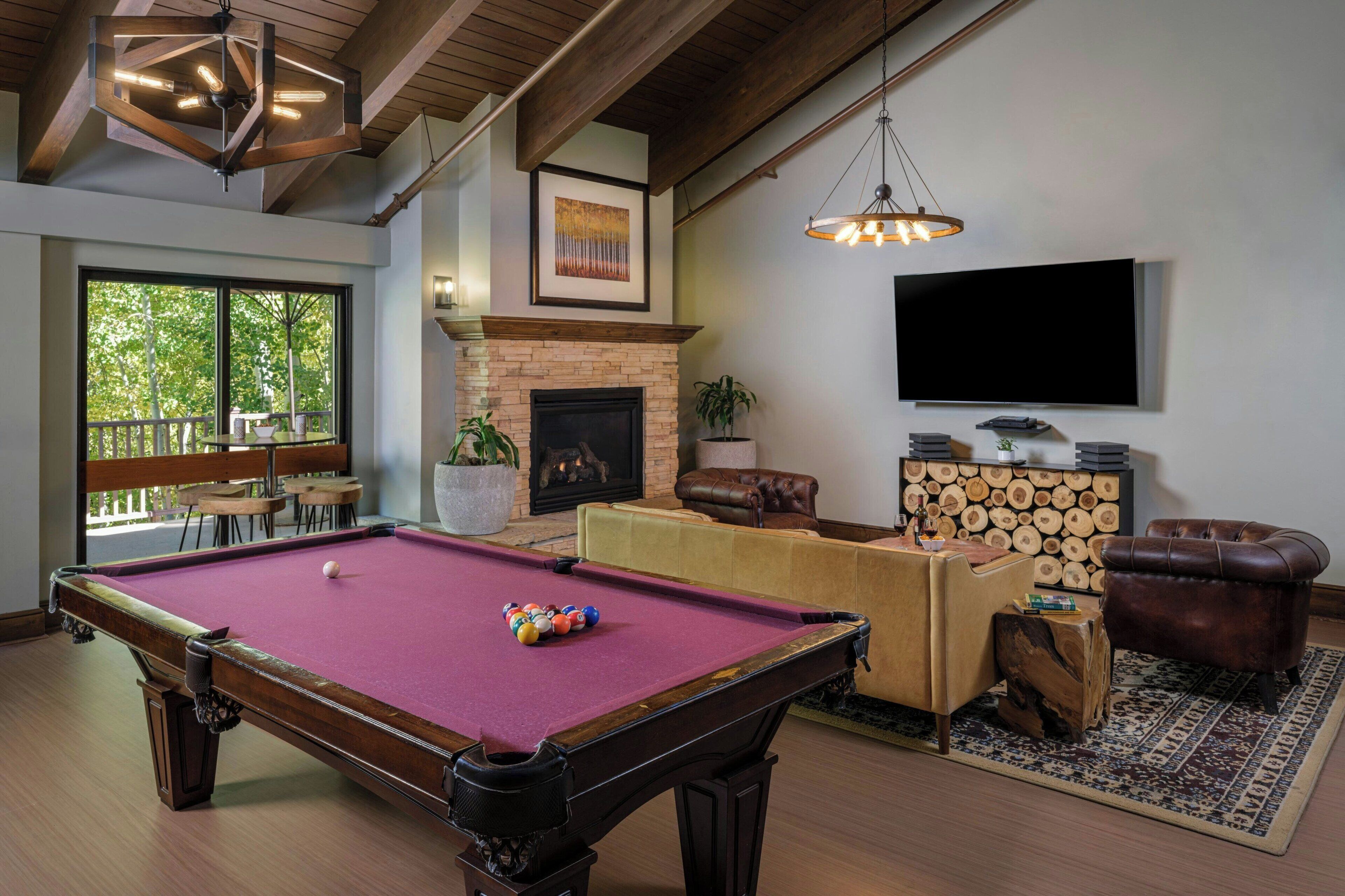 A pool table with balls, a fireplace, a television, and leather furniture in a room with wooden beams and large windows.