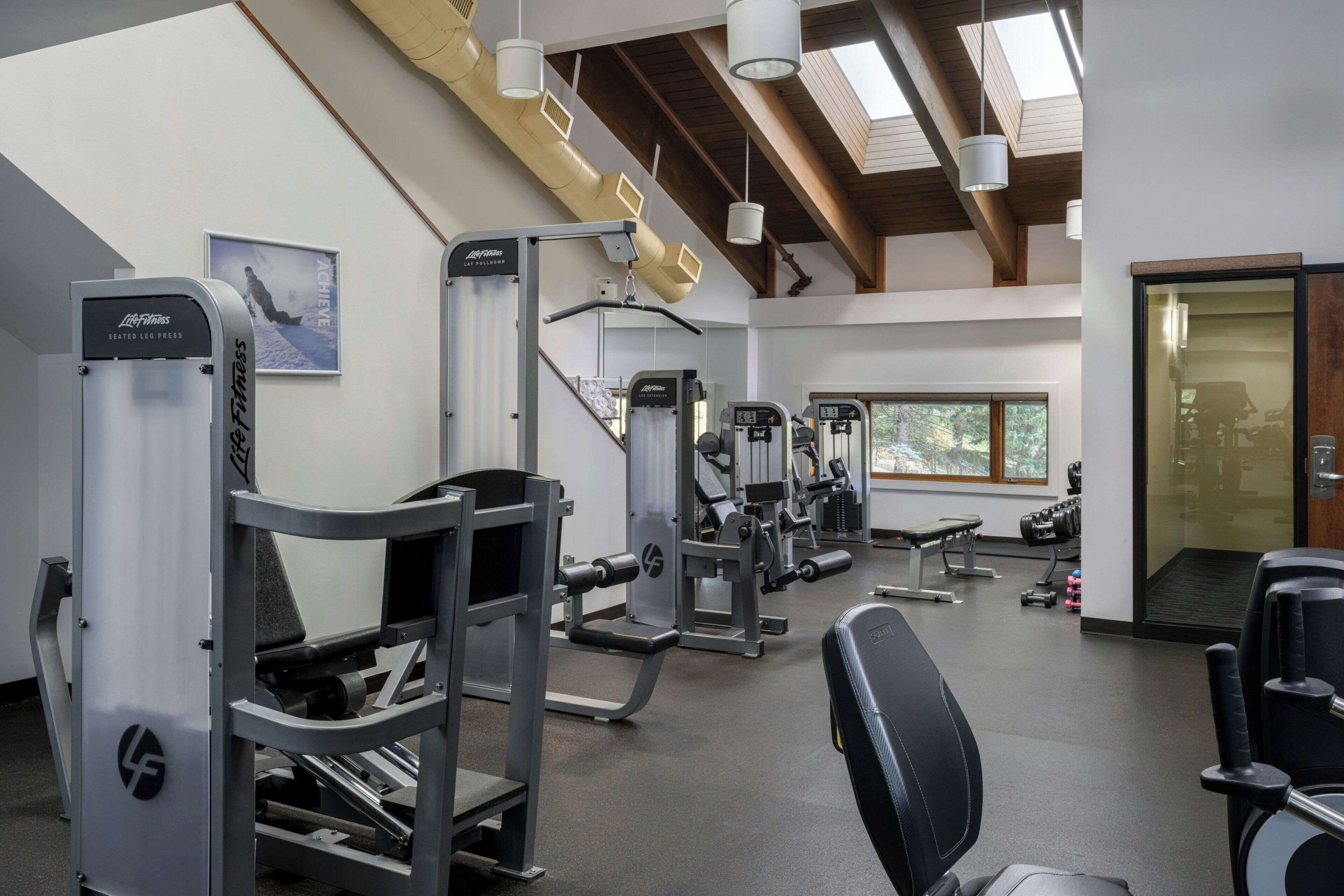 Un gimnasio con diversas máquinas de ejercicio, una banca y un amplio ventanal que proporciona luz natural.