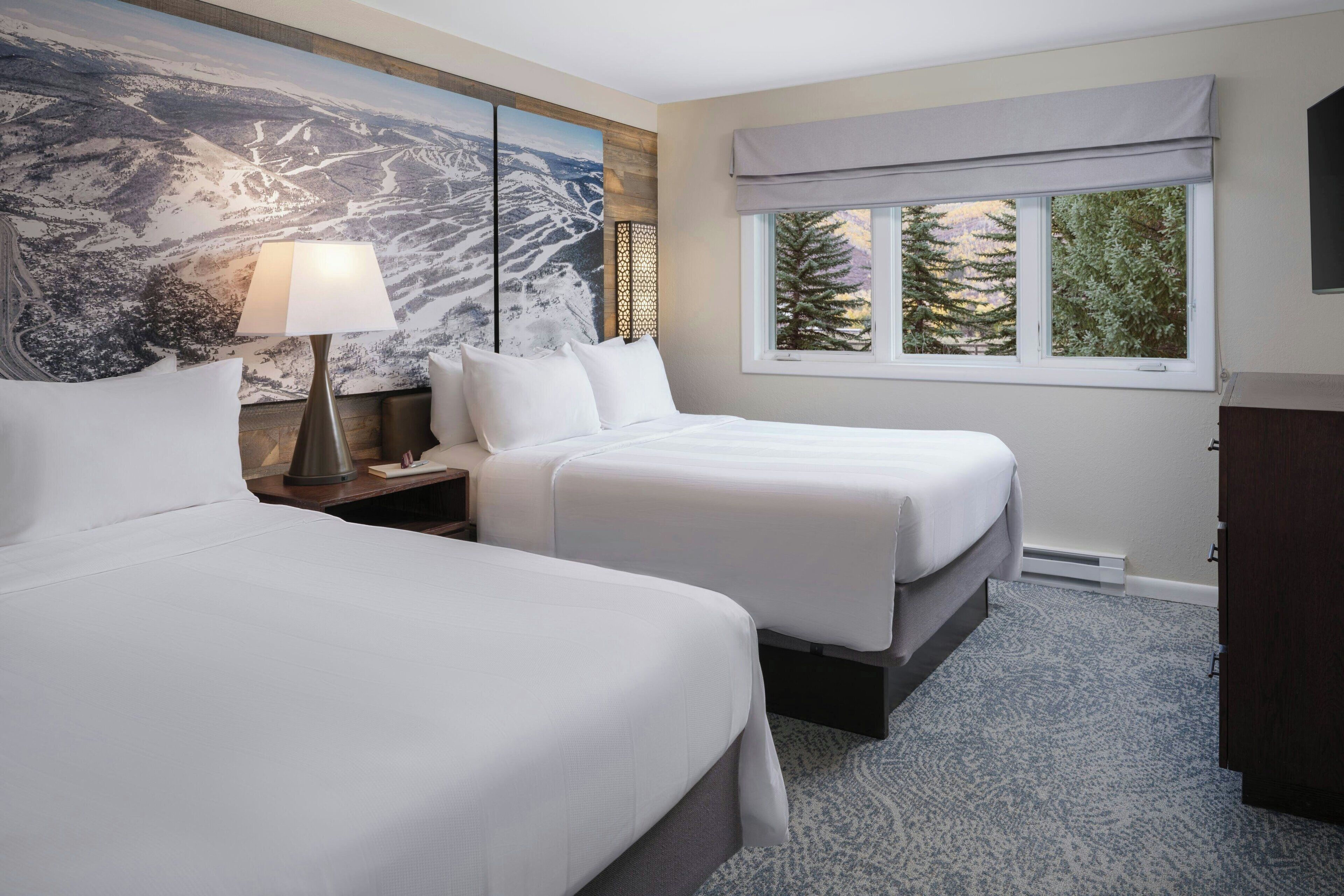 A hotel room with two beds, a large painting on the wall, a window with a view of trees, and a lamp on the nightstand.