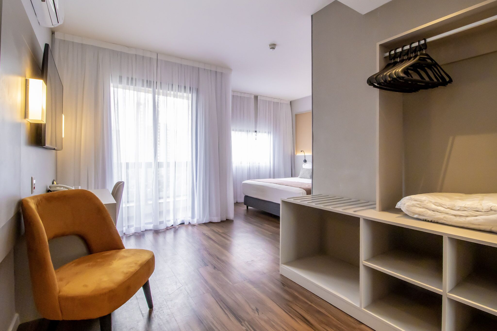 A hotel room with a bed, a chair, a wardrobe, and a desk with a lamp.