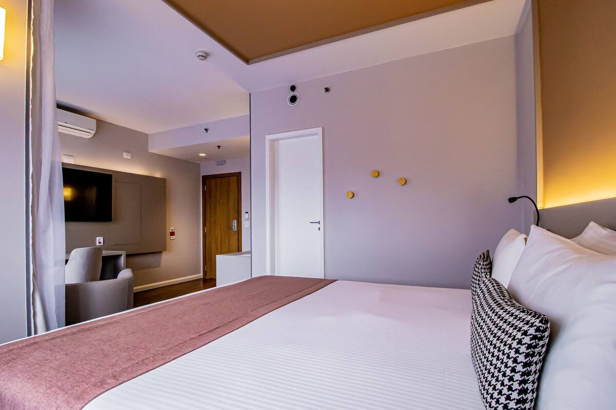 A hotel room with a bed, a desk, a chair, a TV, and a door.