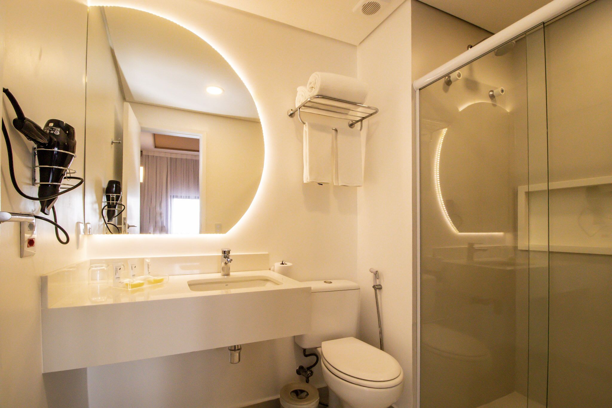 Un baño moderno con lavamanos, espejo y retrete blancos. Cuenta con una cabina de ducha de vidrio y un perchero con toallas.