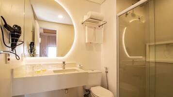 Un baño moderno con lavamanos, espejo y retrete blancos. Cuenta con una cabina de ducha de vidrio y un perchero con toallas.