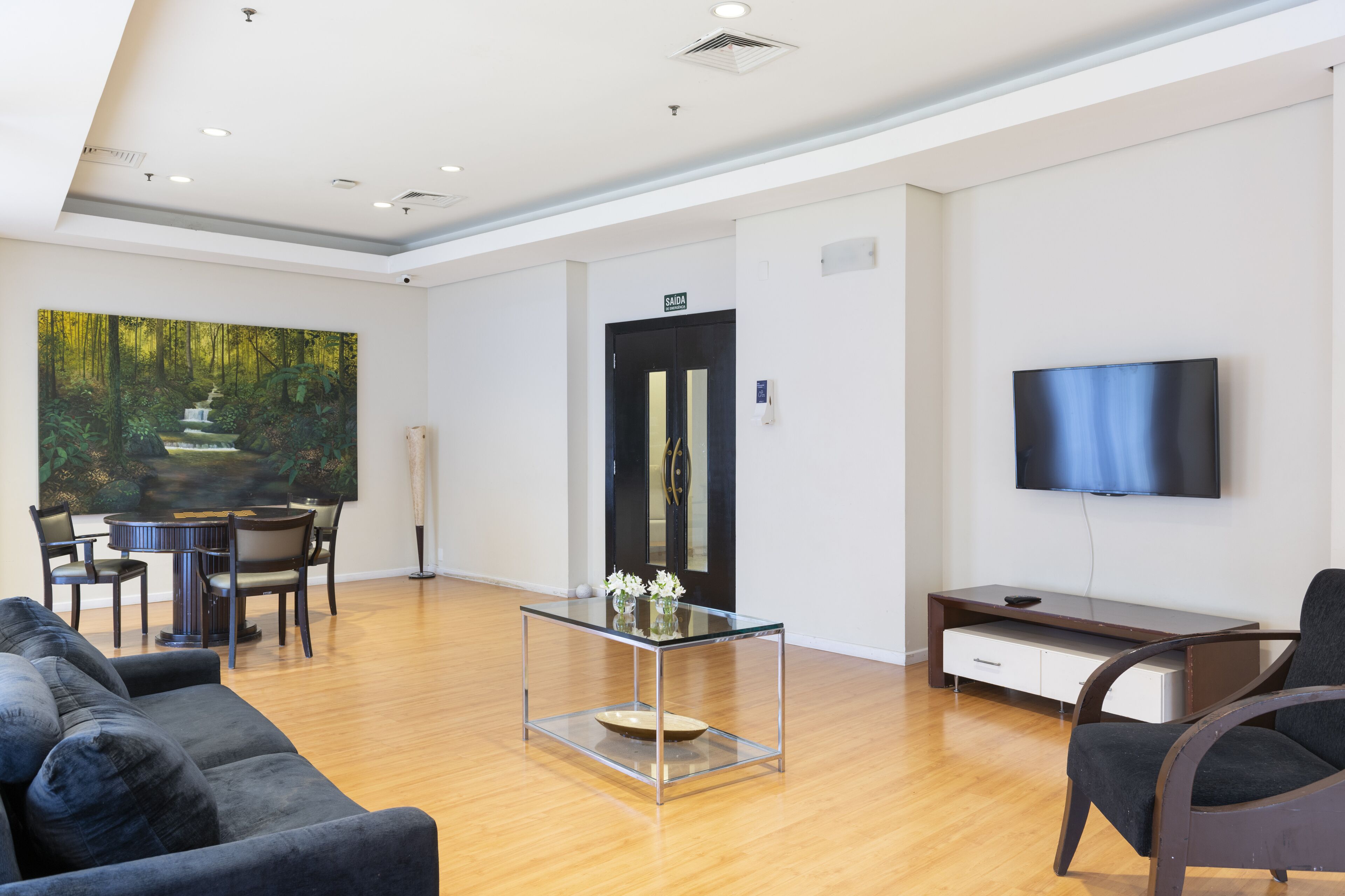 Een moderne hotellobby met een eetgedeelte, een bank, een televisie en een schilderij van een boslandschap.