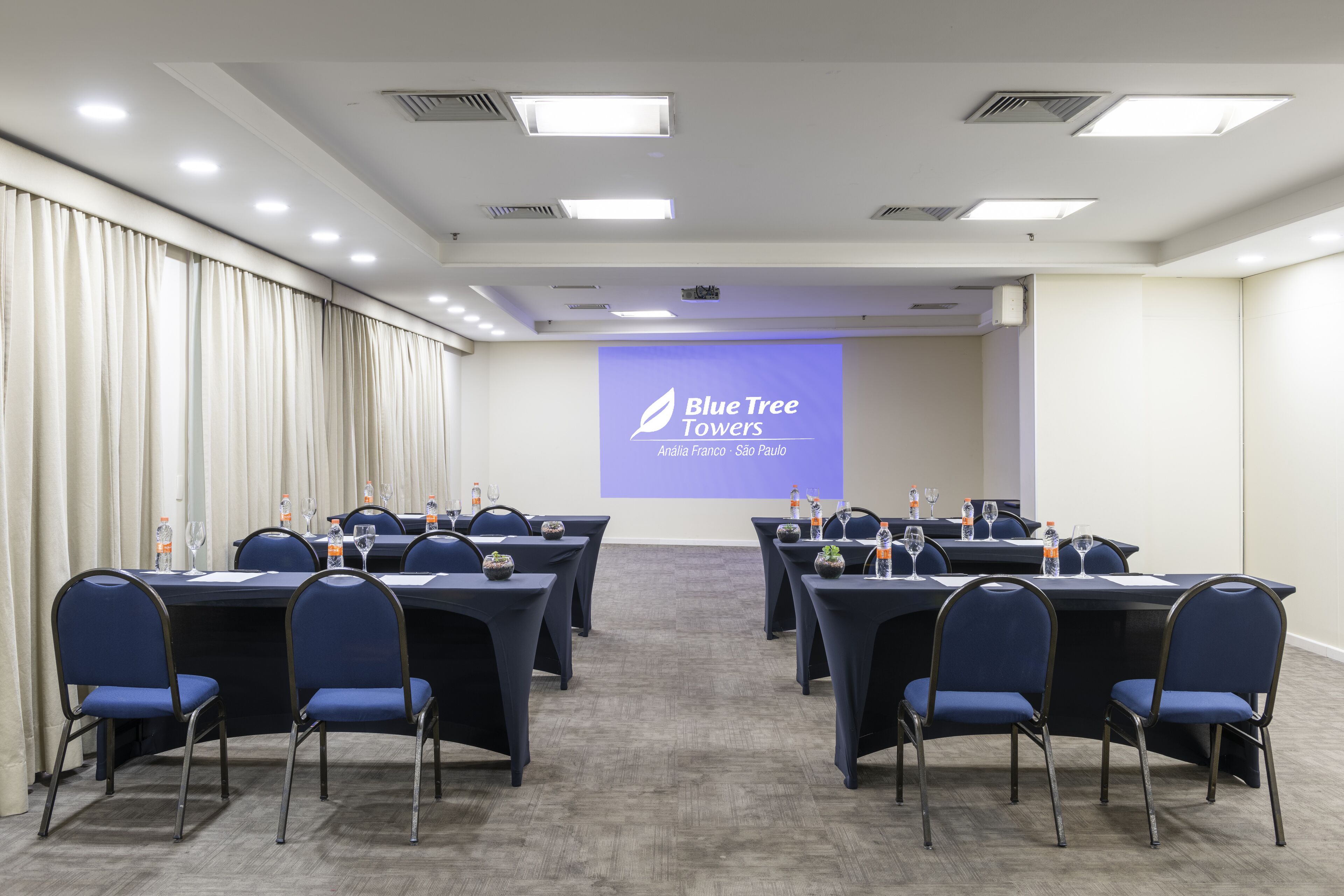 A conference room with tables and chairs, a projector screen displaying "Blue Tree Towers Ana Lila Franco - Sao Paulo", and a white curtain on the left.