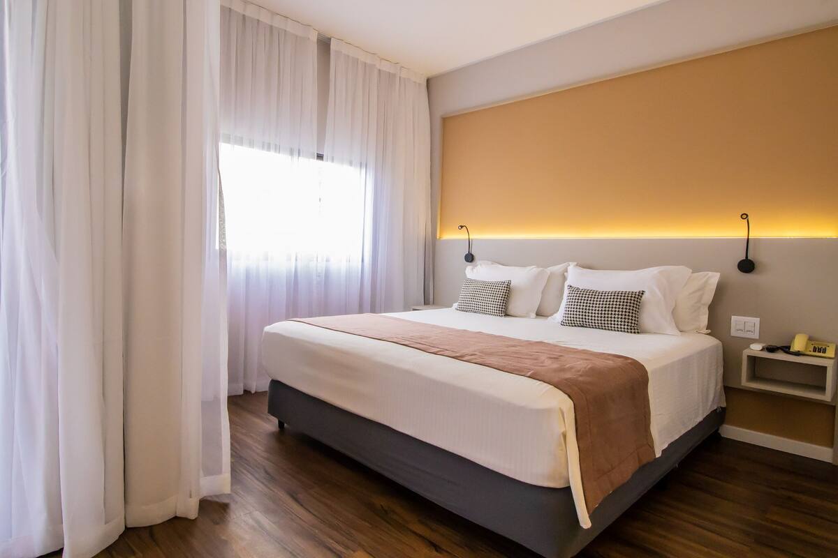 A hotel room with a large bed, white curtains, a bedside table with a lamp, and a wall-mounted shelf.