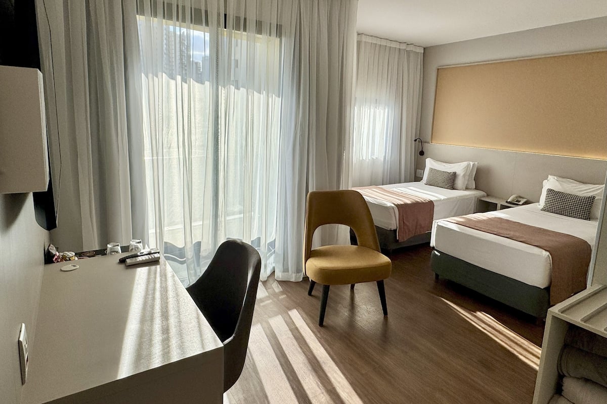 A hotel room with a desk, two beds, a chair, and a large window with sheer curtains.