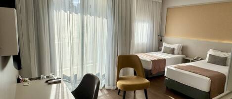 Habitación de hotel con escritorio, dos camas, una silla y un ventanal con cortinas ligeras.